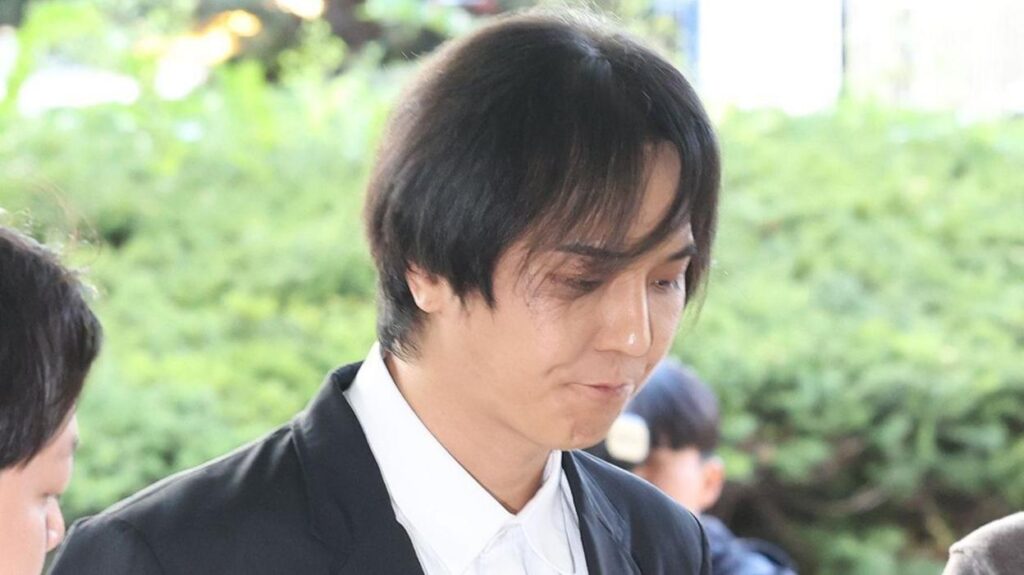 Song Min-ho in a dark blazer and white buttoned shirt looks down as he is surrounded by reporters.