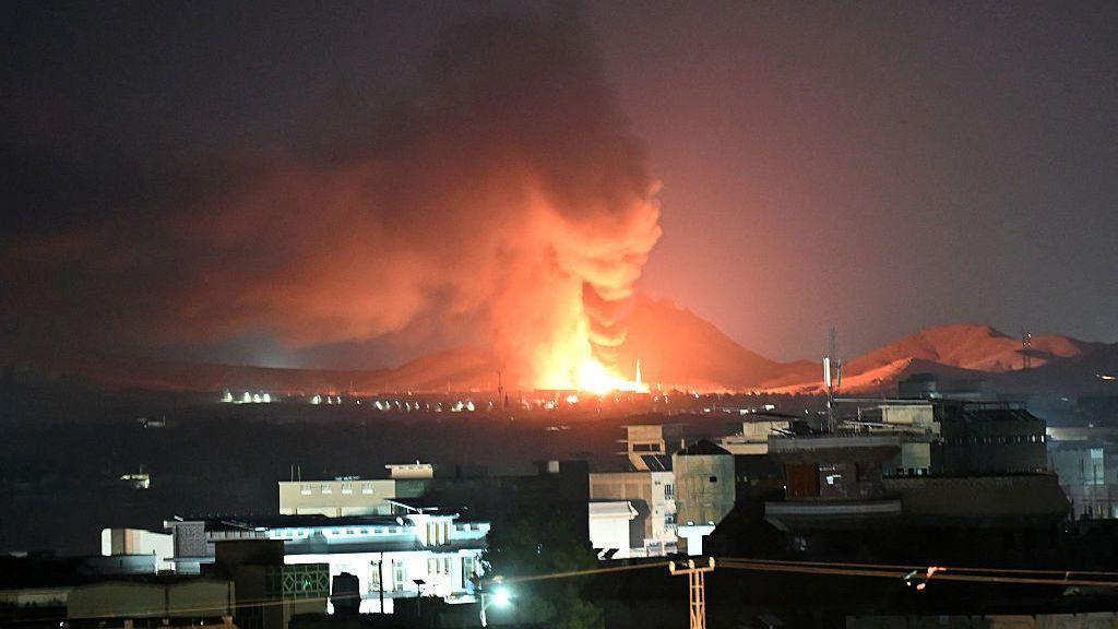 A large fireball and thick column of smoke rise into the night sky on the outskirts of a city, with buildings and hills visible nearby.