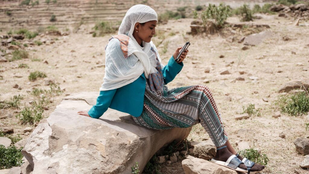 A woman sits on a rock in an arid landscape looking at her phone. She is wearing a white headscarf, blue jacket and a patterned skirt