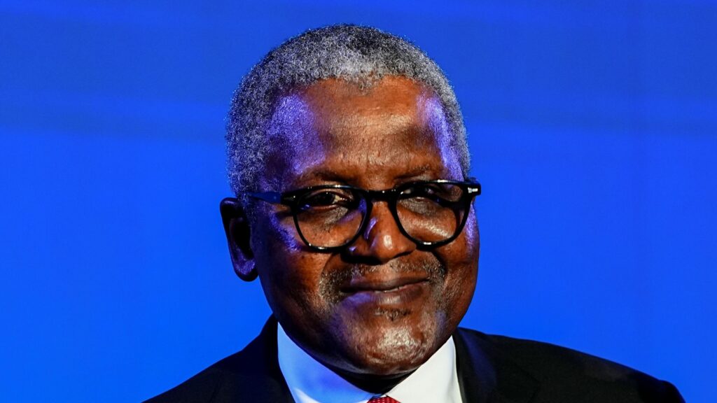 A man with grey hair, wearing glasses with black frames, a black suit, a white shirt, and a red tie, smiles into the camera