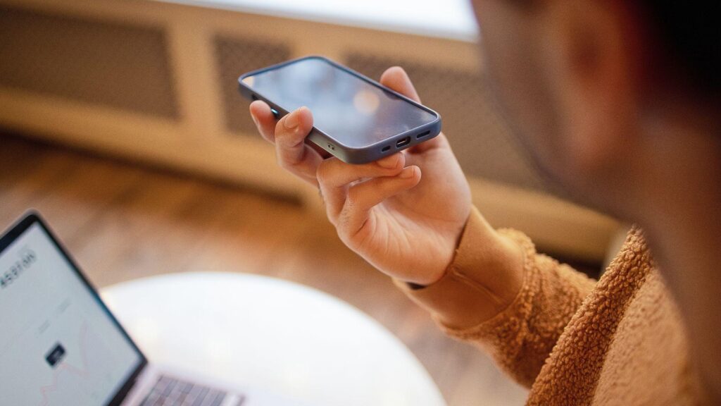 Stock photo shows a person speaking into a phone while sitting at a desk indoors on their laptop.