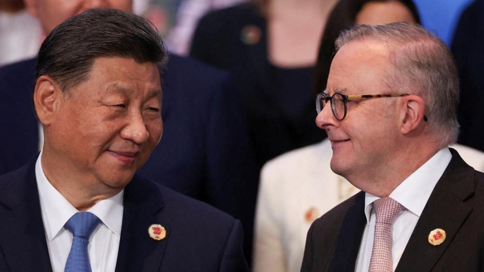 Two older men dressed in suits and ties smiles at each other