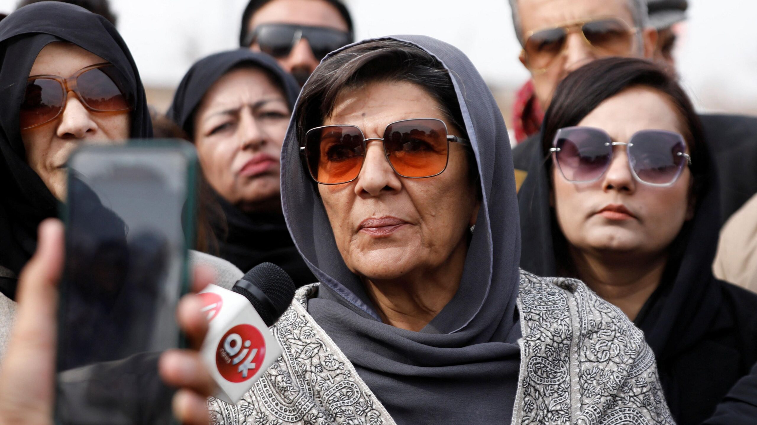 Aleema Khanum wears a grey head covering and sunglasses as she speaks into a news microphone. There are other women standing behind her.