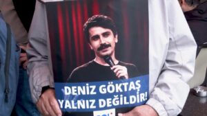 A poster of Deniz Göktaş, who has dark short hair and a moustache, holding a microphone with a red curtain behind him. The words "Deniz Göktaş is not alone!" are written on the poster.