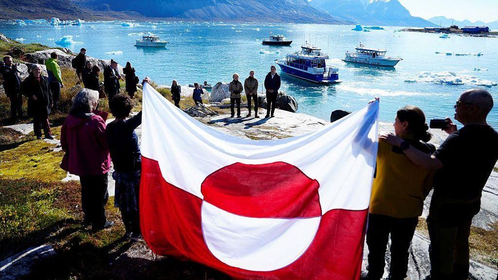 People hold a flag on the shoreline of Bear Island with boats in the background