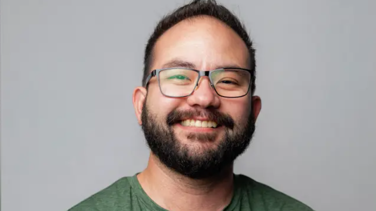 Jonathan McKinsey seen with a white background, smiling, wearing glasses and a beard and green shirt