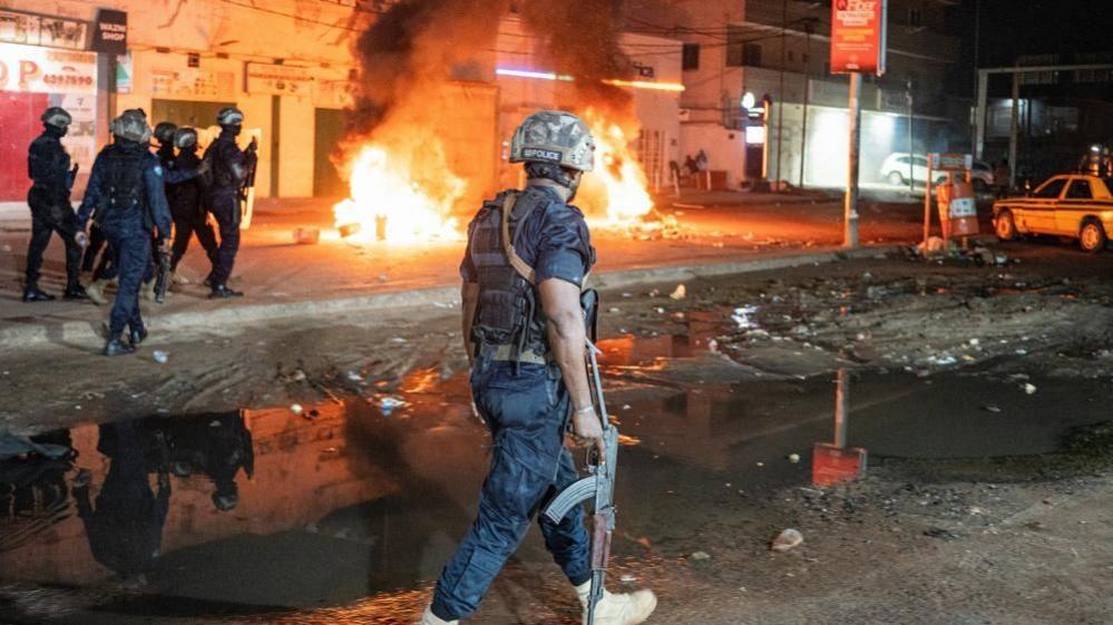 Police officers patrol a street as burning tyres and debris send flames and thick smoke into the air during protests over prolonged power cuts in The Gambia.