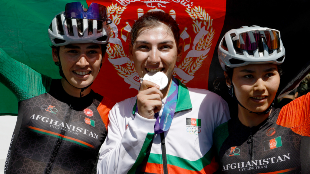 Fariba Hashimi pretends to bite her medal after winning the Aichi-Nagoya Asian Games women's cycling road race silver in Shinshiro, as she stands between two Afghanistan fellow riders