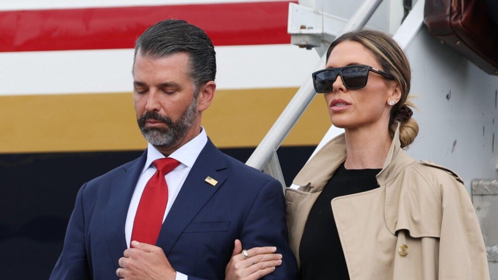 Donald Trump Jr. and his wife Bettina Anderson are seen close up with a plane behind them. She is on the right in a blacktop, tan trench coat and black sunglasses. Her hair is pulled back. He is wearing a blue suit and red tie.