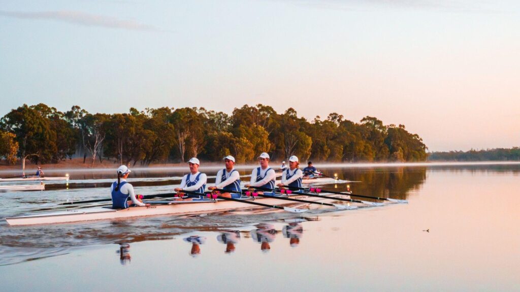 A rowing boat with rowers wearing white and blue row down a wide river with mist rising from the water.