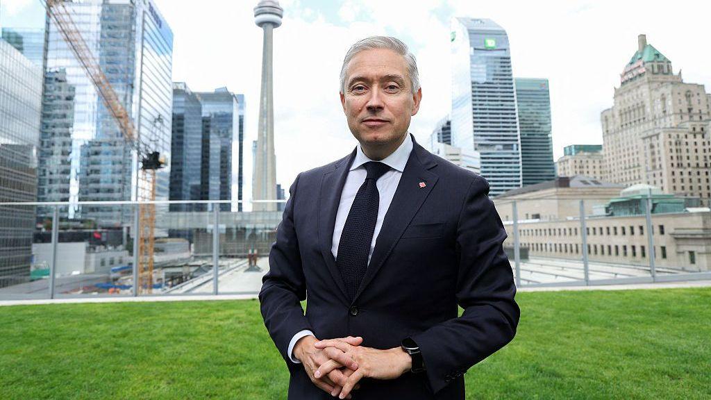 Champagne wears a suit and tie while he stands on a lawn in front of the Toronto city skyline of high-rise buildings.