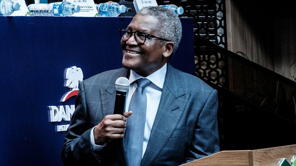 Aliko Dangote, wearing a suit, is smiling as he turns to the left of the picture. He is holding a microphone in his right hand.