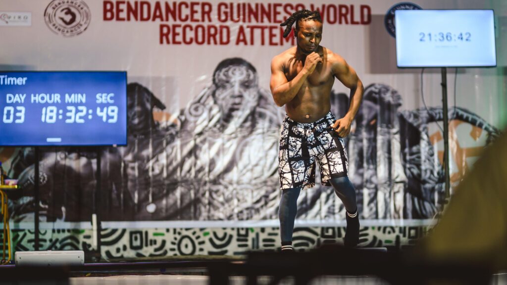 Ben Dancer dancing on a stage surrounded by supporters. Behind him a screen showing how many days he danced non-stop.