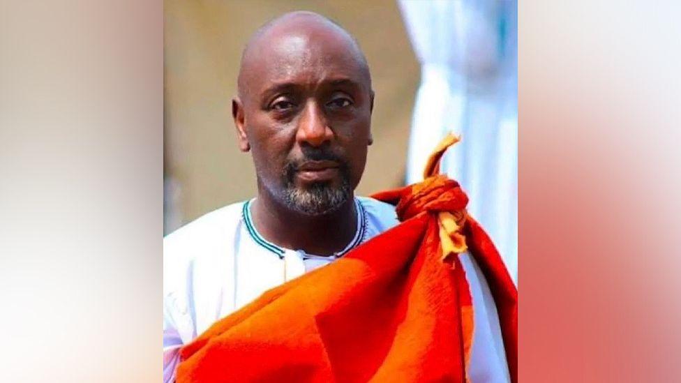 Edward Rukidi Kijanangoma wearing traditional clothing of a white gown with an orange cloth tied around the neck