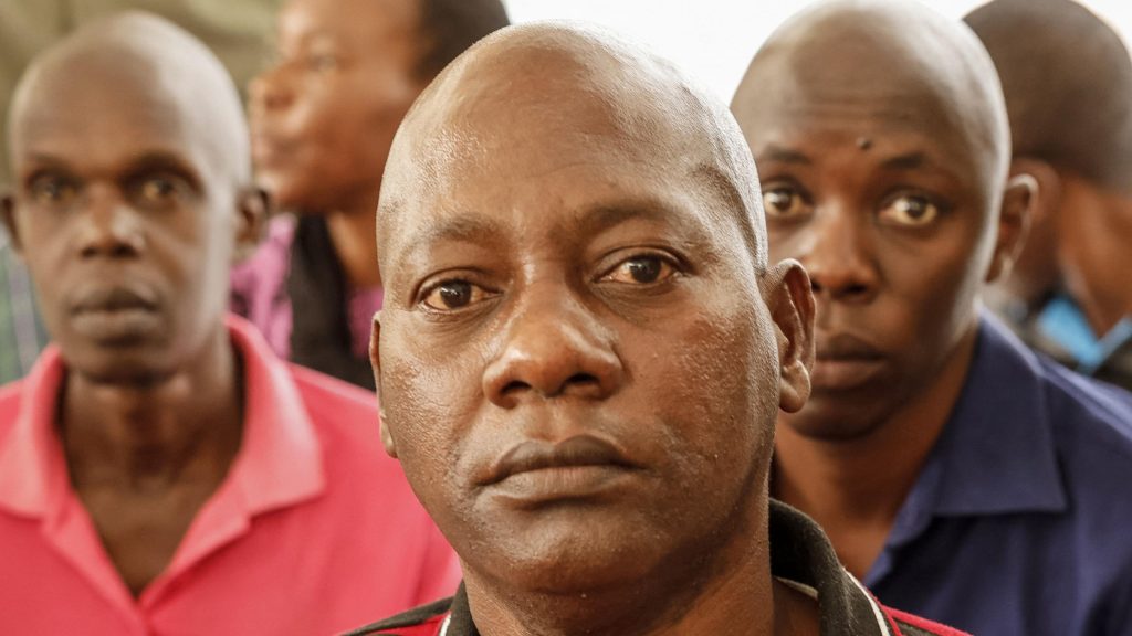 A close shot of a bald man staring at the camera. Two other men can be seen behind him