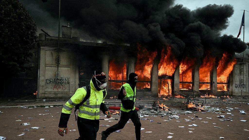 Protesters wearing yellow vests run past Lyon's tourism office set on fire during a demonstration to protest against the "high cost of living", in Lyon, southeastern France, on October 17, 2026