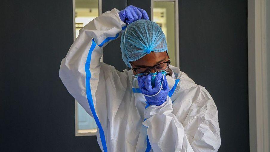 A health worker, wearing full protective clothing and gloves, puts on a face mask