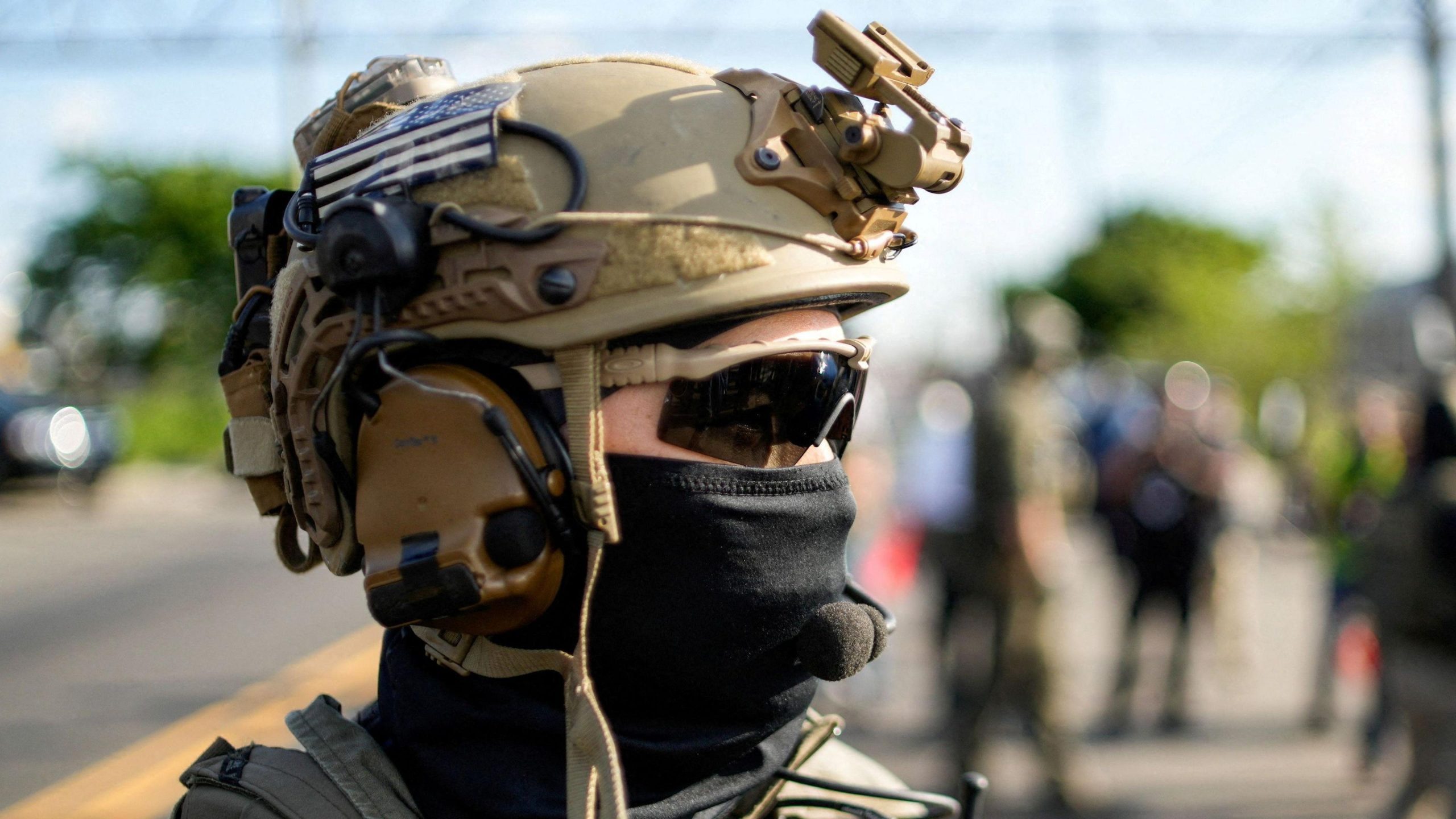 a US Immigration and Customs Enforcement officer standing outside of a detention center in New Jersey wearing a tactical helmet, wrap-around sunglasses, and a cloth covering most of his face.