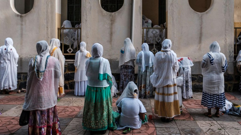 Women wearing colourful traditional Ethiopian dresses in green, red and gold, with long headscarves, pray outside a church. Some stand while others kneel.