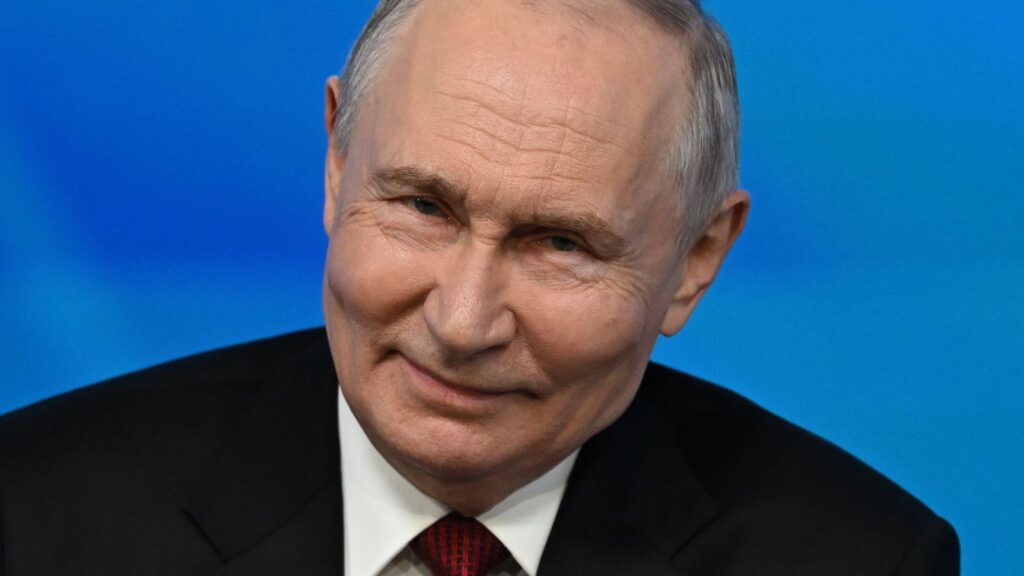 Close up of Vladimir Putin while speaking to an audience at the Valdai foreign policy forum. He has short grey hair and is wearing a black suit, white shirt and red tie.