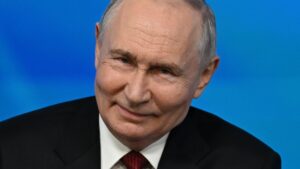 Close up of Vladimir Putin while speaking to an audience at the Valdai foreign policy forum. He has short grey hair and is wearing a black suit, white shirt and red tie.