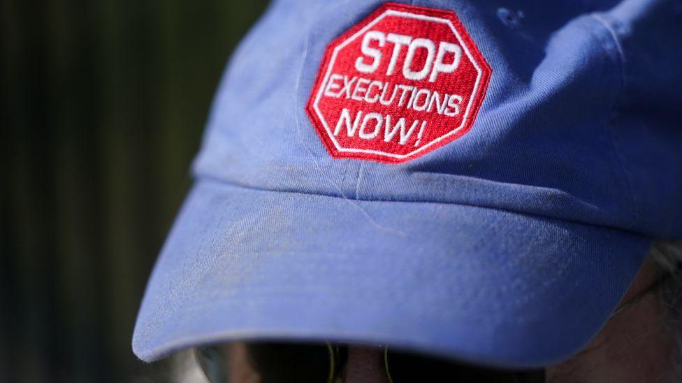 Blue baseball cap with the words STOP EXECUTIONS NOW! written on the front in white letters on a red badge. You cannot see the face of the person wearing it, only their sunglasses