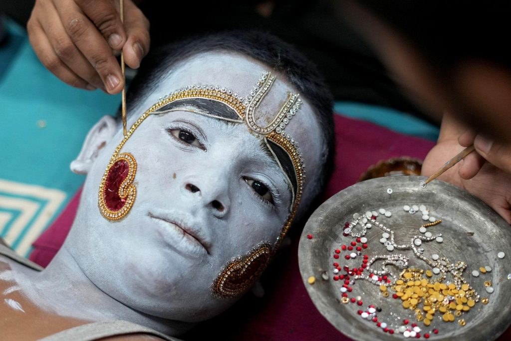A person with their face painted a pale blue with ornate blue and gold jewellery across their face looks towards the camera while another person applies small jewelled studs from a dish, in Uttar Pradesh on Sunday.