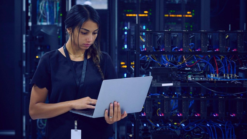 A female technician dressed in navy blue clothes and wearing a lanyard around her neck is standing in a server room using her laptop.