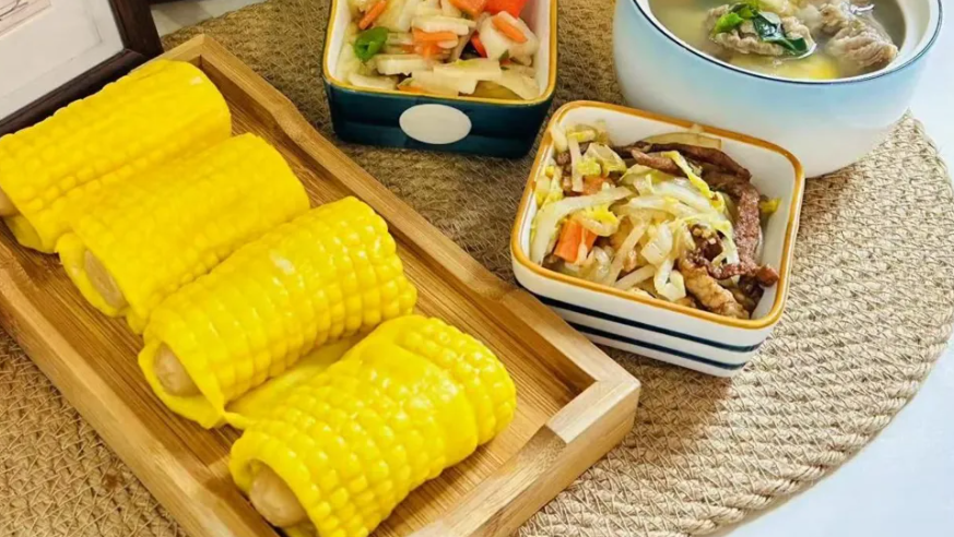 A photo of one of the meals offered by the nursery. On a wooden tray are four very bright yellow sausage corn buns. Three other plates positioned next to it are an assortment of vegetables and meat, and a bowl of soup