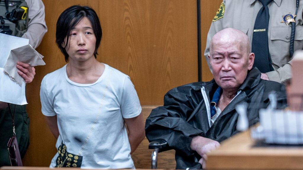 Silvia Zhang, left, and Guojun Xuan, right, sitting in Pasadena courtroom with two sheriffs standing behind them. Zhang has her hand behind her back and is wearing a blue shirt while Xuan is wearing a black jacket