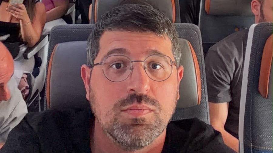 A selfie of Adam Polo sitting on a busy aeroplane with a grey and orange seat cushion. He has short dark hair and a beard and is wearing glasses and a black t-shirt