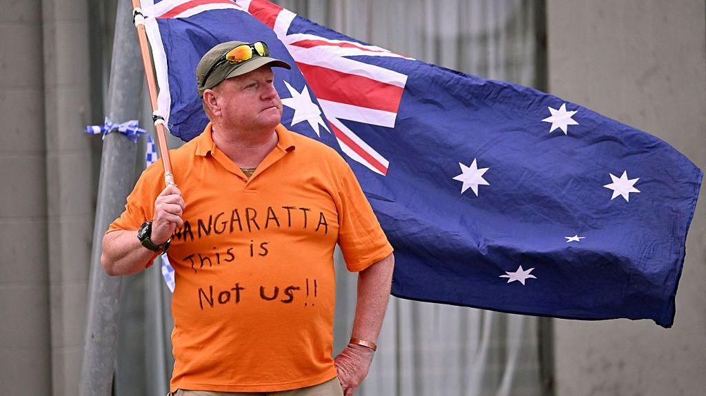 A man wearing an orange t-shirt reading "Wangaratta this is not us!!" waves an Australian flag