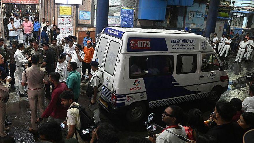 An ambulance carrying Durga Puja Pandal stampede victims stops in front of a hospital. Lots of men are seen standing around the white vehicle.