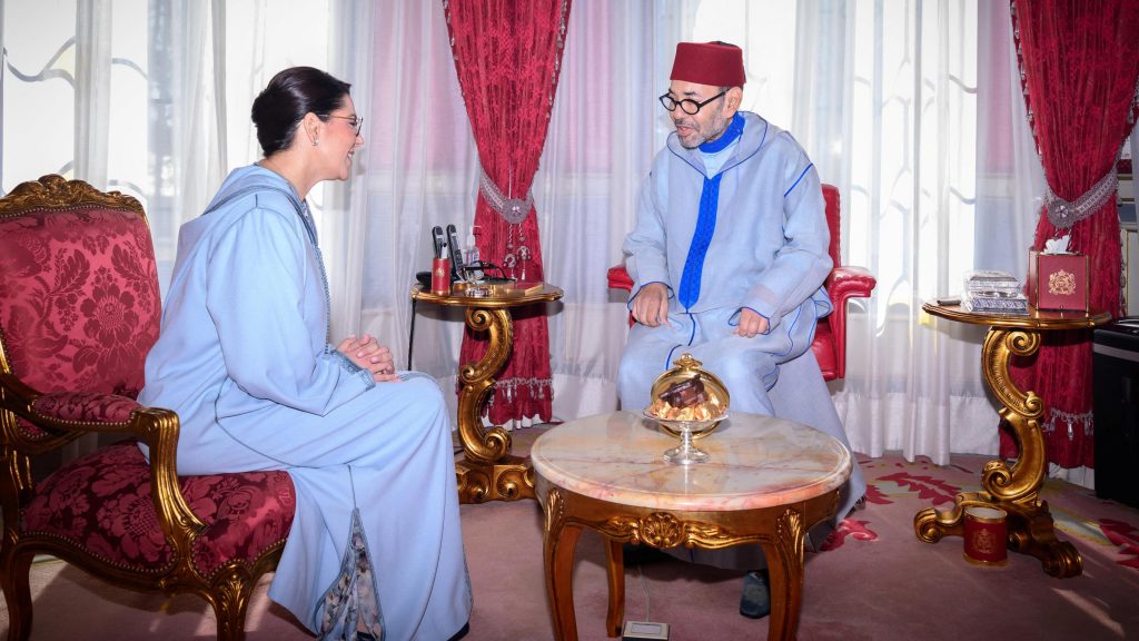 At the royal palace, El Mansouri, dressed in a long blue traditional robe, sits on a red-and-gold chair facing King Mohammed VI. He is wearing a long blue traditional robe and a red Moroccan hat.