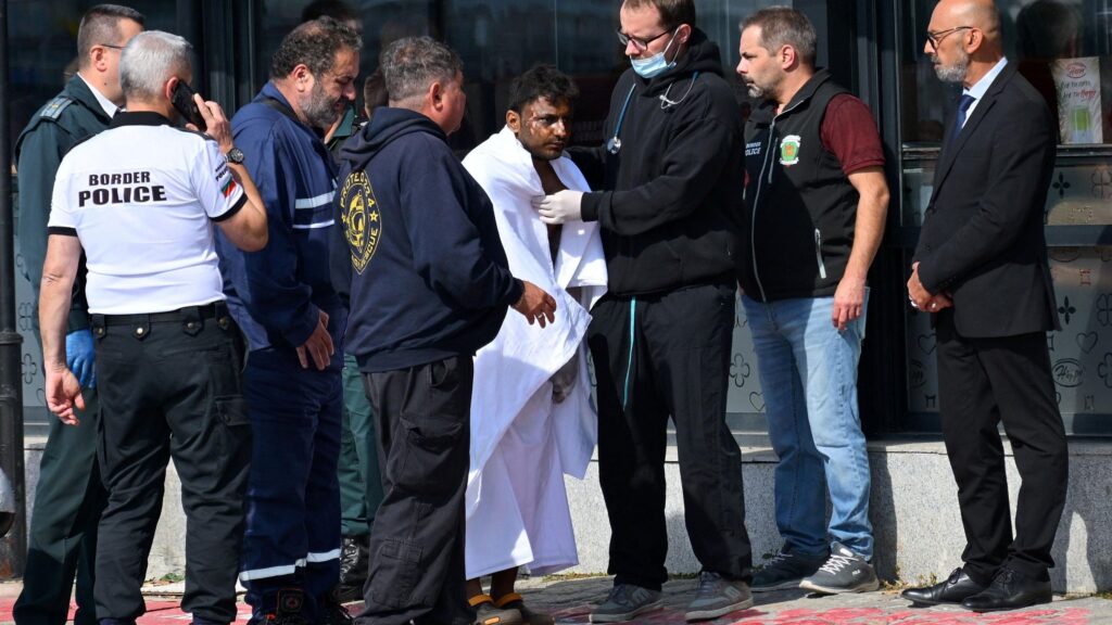 A man is wrapped in a large white blanket after being rescued from an attacked ship. There are seven people around him, one of whom has a stethoscope around his neck