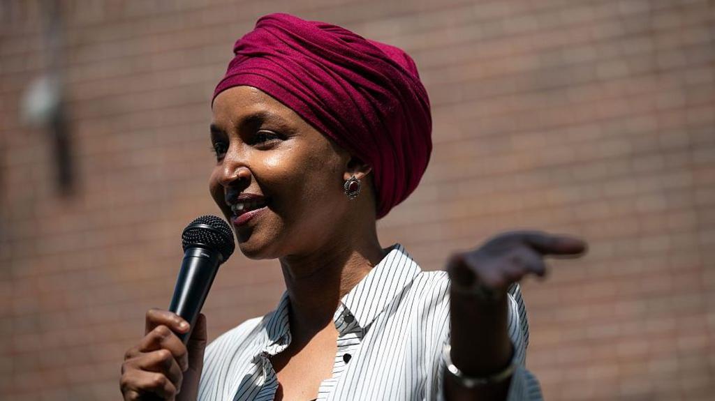 Omar in magenta turban and black and white shirt with thin dark stripes, holds out her left hand while her right one holds a black microphone