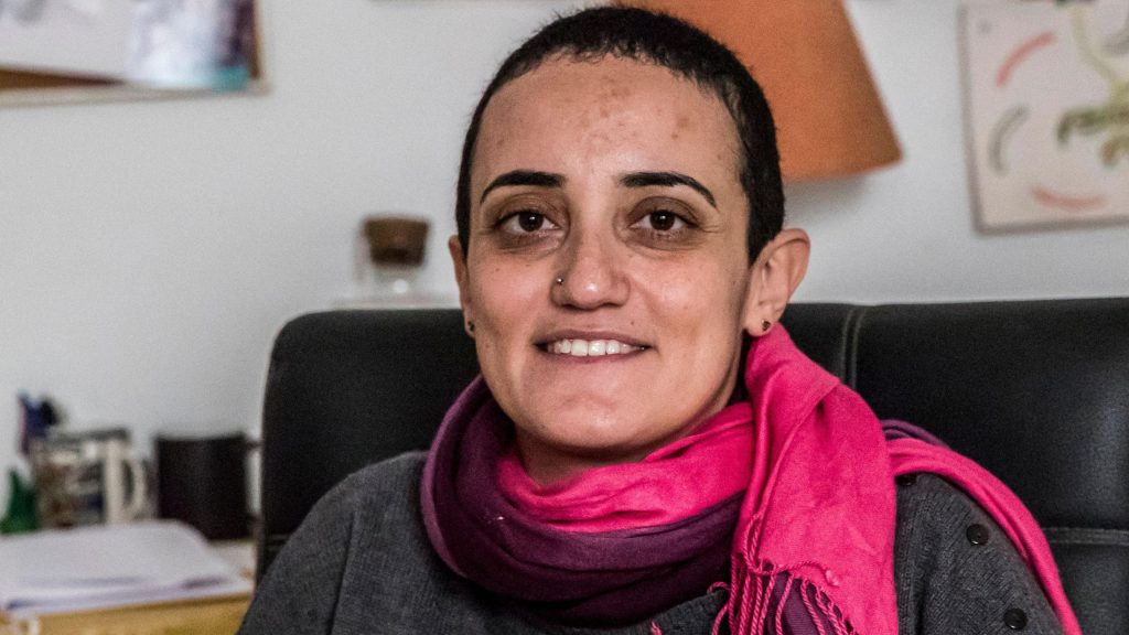 Egyptian journalist Lina Attalah sits on a black leather chair. She has a red scarf around her neck, a grey top, cropped black hair, stud earrings and and a silver metal studd in her right nostril, a broad smile on her lips.
